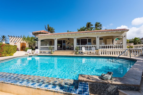 A poolside view of a resort with a clear blue pool, a covered seating area, and a white building with a tiled roof. - Aruban villa in Nord, great view, cozy, safe, short drive to many beaches (Noord)