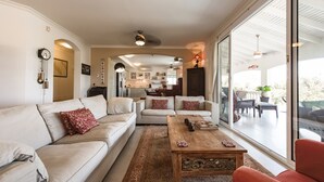 Amplia sala de estar con sofá, mesa de centro y vistas a la cocina y un espacio exterior con mobiliario.