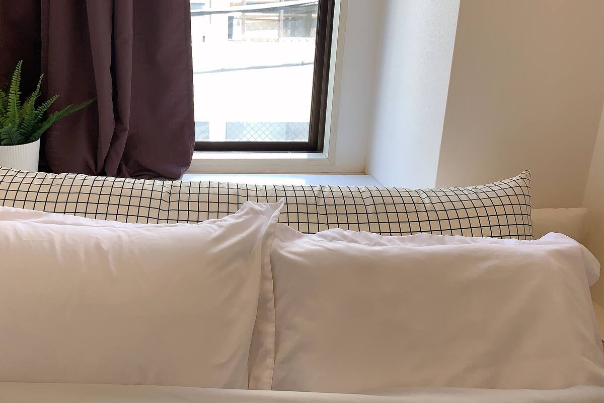A white sofa with patterned cushions, a window with a view of buildings, and a potted plant on the left.
