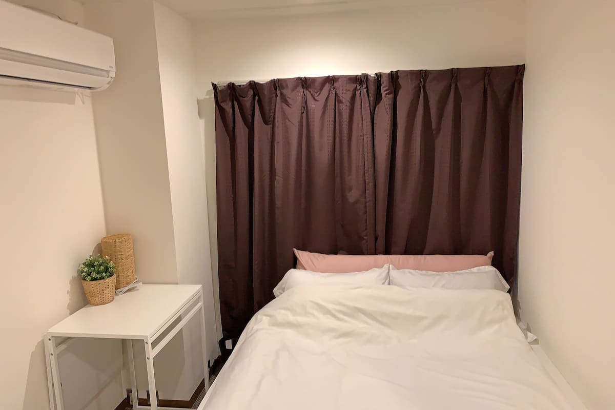 A bedroom with a bed, a white bedside table, a wicker basket, and a brown curtain.
