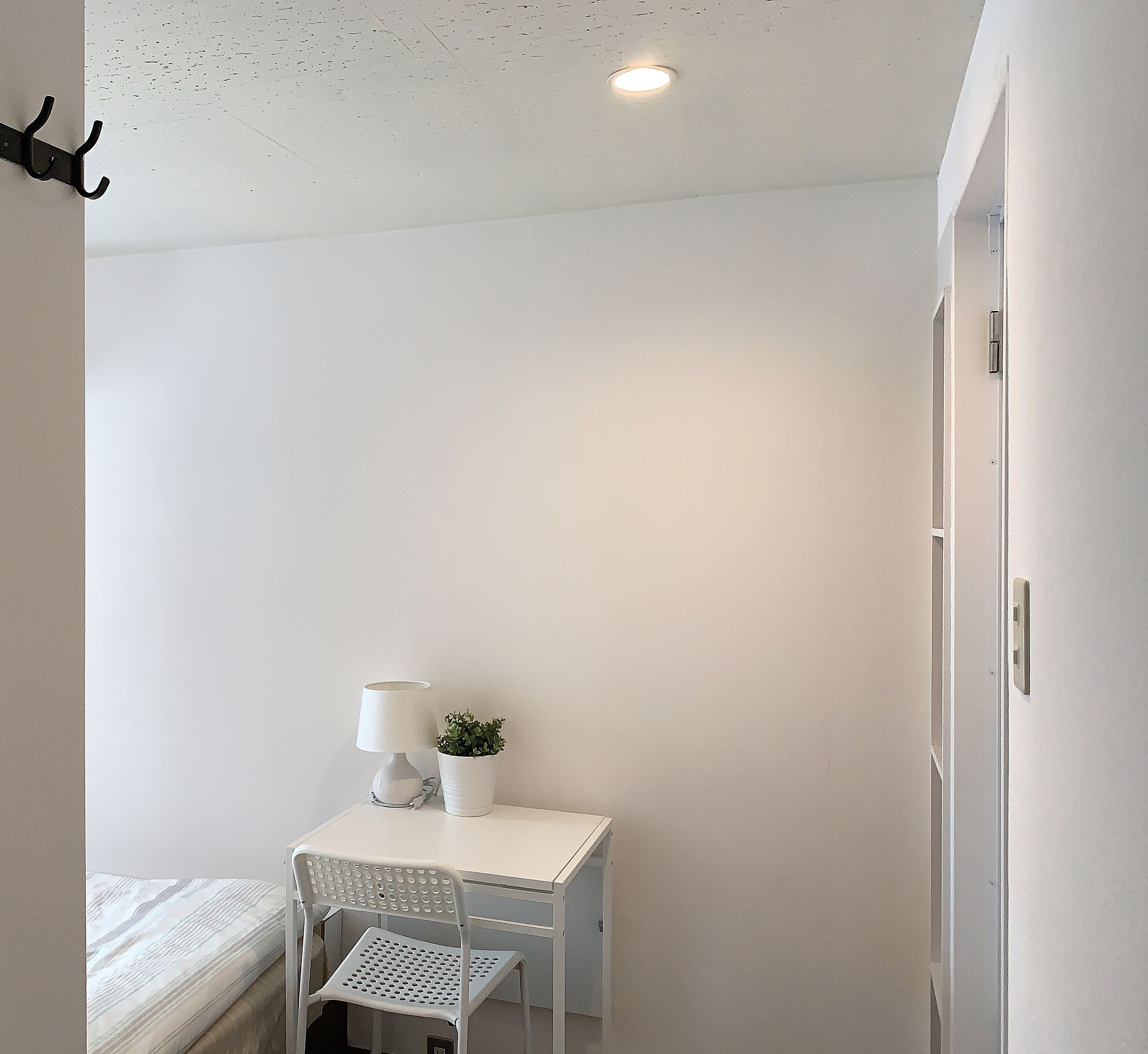 A small, white room with a single bed, a white desk with a lamp and a small plant, and a white chair.