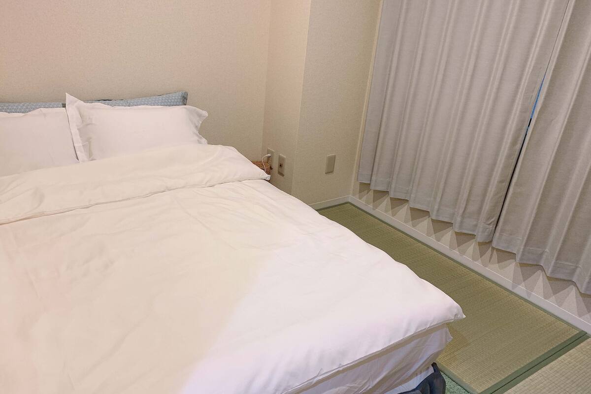 A neatly made bed with white linens, a small bedside table with a lamp, and a window with curtains.