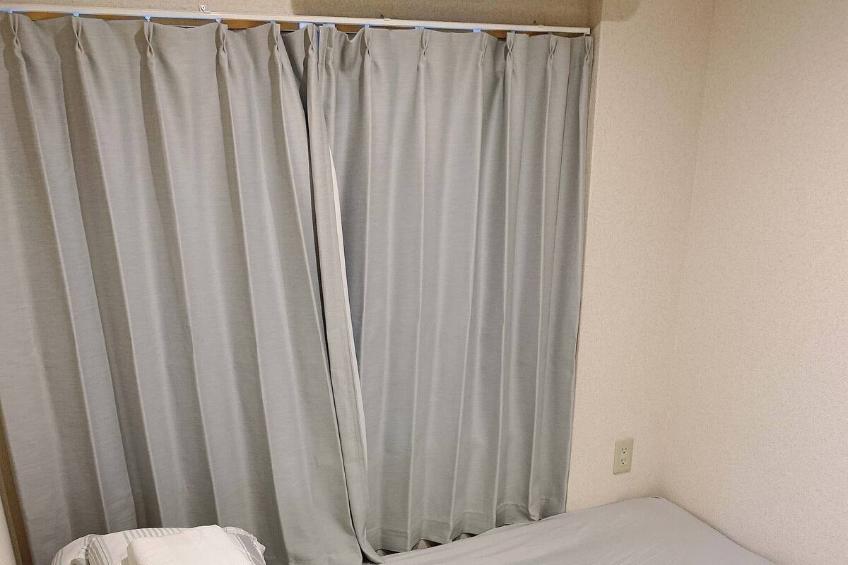 A single bed with a grey mattress and striped pillows, a grey curtain, and a plain wall with a power outlet.