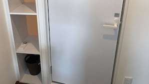 A closed door with a white handle, a black trash bin, and a white shelf unit with multiple compartments. - Hotel Minami (Tokyo)