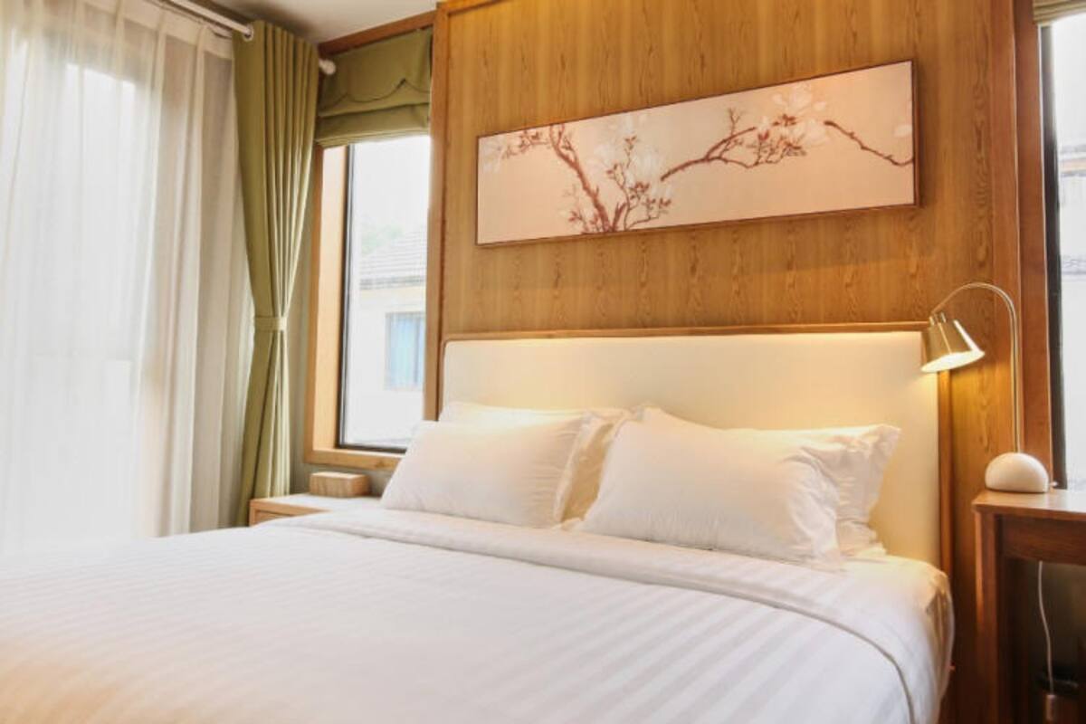 A neatly made bed in a hotel room with a wooden headboard, a nightstand with a lamp, and a window with curtains.