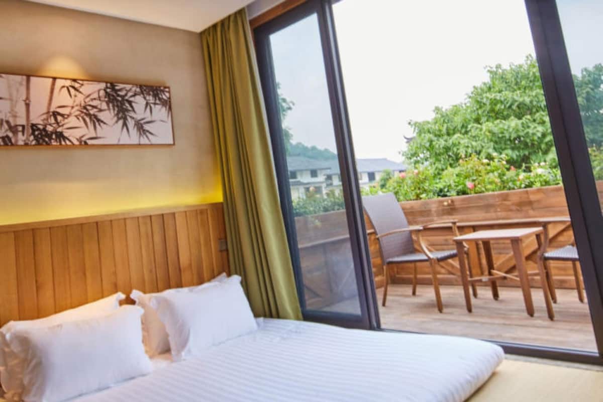 A hotel room with a large bed, a wooden headboard, a window with a view of greenery, and a balcony with a table and chairs.