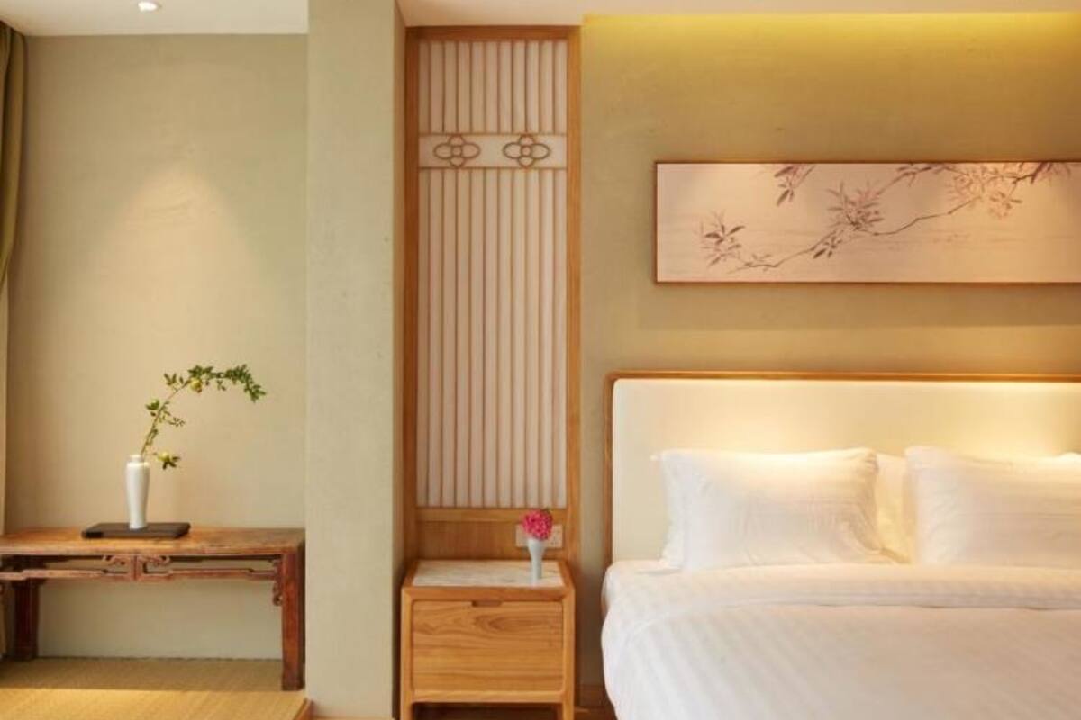 A hotel room with a large bed, a bedside table, a small wooden nightstand, a vase with flowers, and a wall-mounted artwork.