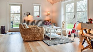 Een gezellige woonkamer met een rieten bank, een witte salontafel en uitzicht op een tuin door grote ramen.