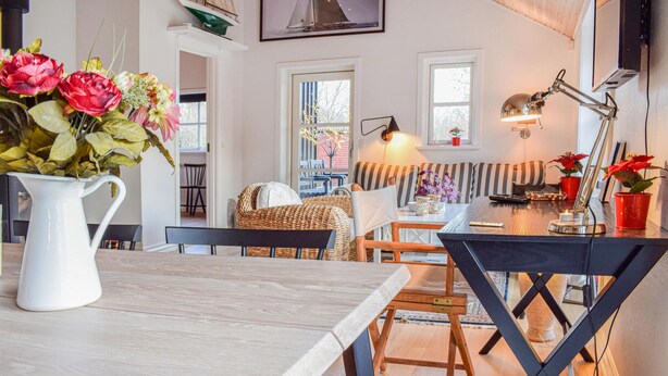 Een gezellig eetgedeelte met een houten tafel, stoelen en een bloevas. Er is een raam met uitzicht naar buiten, een lamp op de tafel en een decoratie van een zeilboot aan de muur.