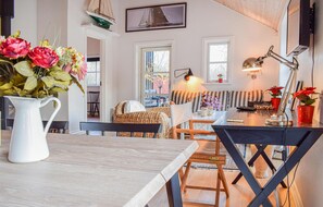 A cozy dining area with a wooden table, chairs, and a vase of flowers. There is a window with a view of the outside, a lamp on the table, and a sailboat decoration on the wall.