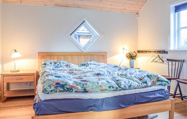 A bedroom with a wooden bed, a nightstand with a lamp, a small window, and a wall-mounted shelf with a vase.
