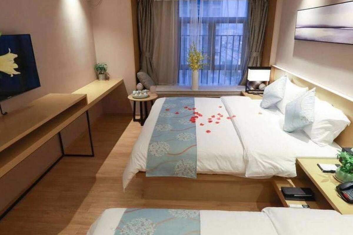 Ein Hotelzimmer mit Bett, Nachttisch, kleinem Schreibtisch, Stuhl, einem Fenster mit Vorhängen und einem an der Wand befestigten Fernseher.