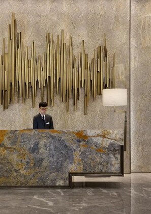 A reception desk with a marble counter and a vertical gold sculpture on the wall. - Oriental Ginza International Hotel (Dongguan)