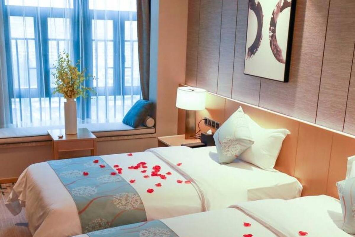 Ein Hotelzimmer mit zwei Betten, einem Fenster mit blauen Vorhängen, einem Nachttisch mit Lampe und einer Vase mit Blumen.