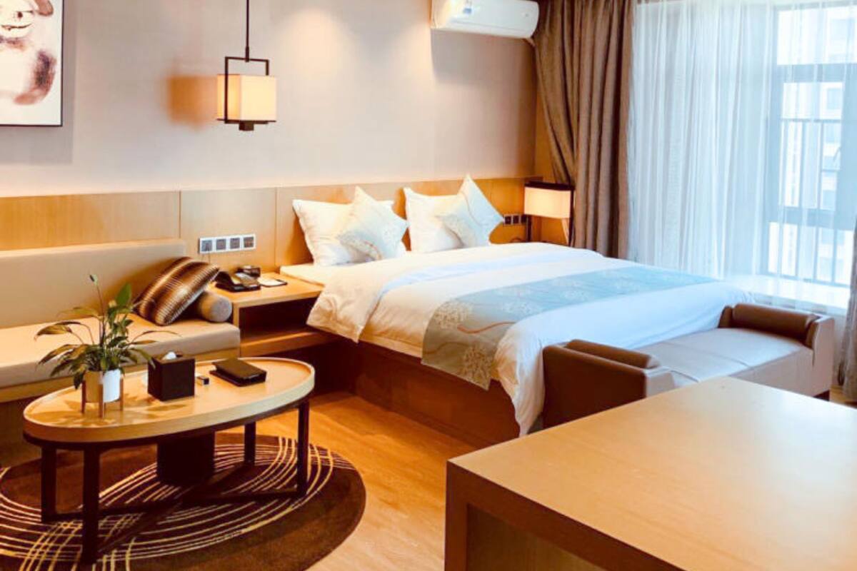 Ein Hotelzimmer mit Bett, Nachttisch, Lampe und Blick ins Freie durch das Fenster.