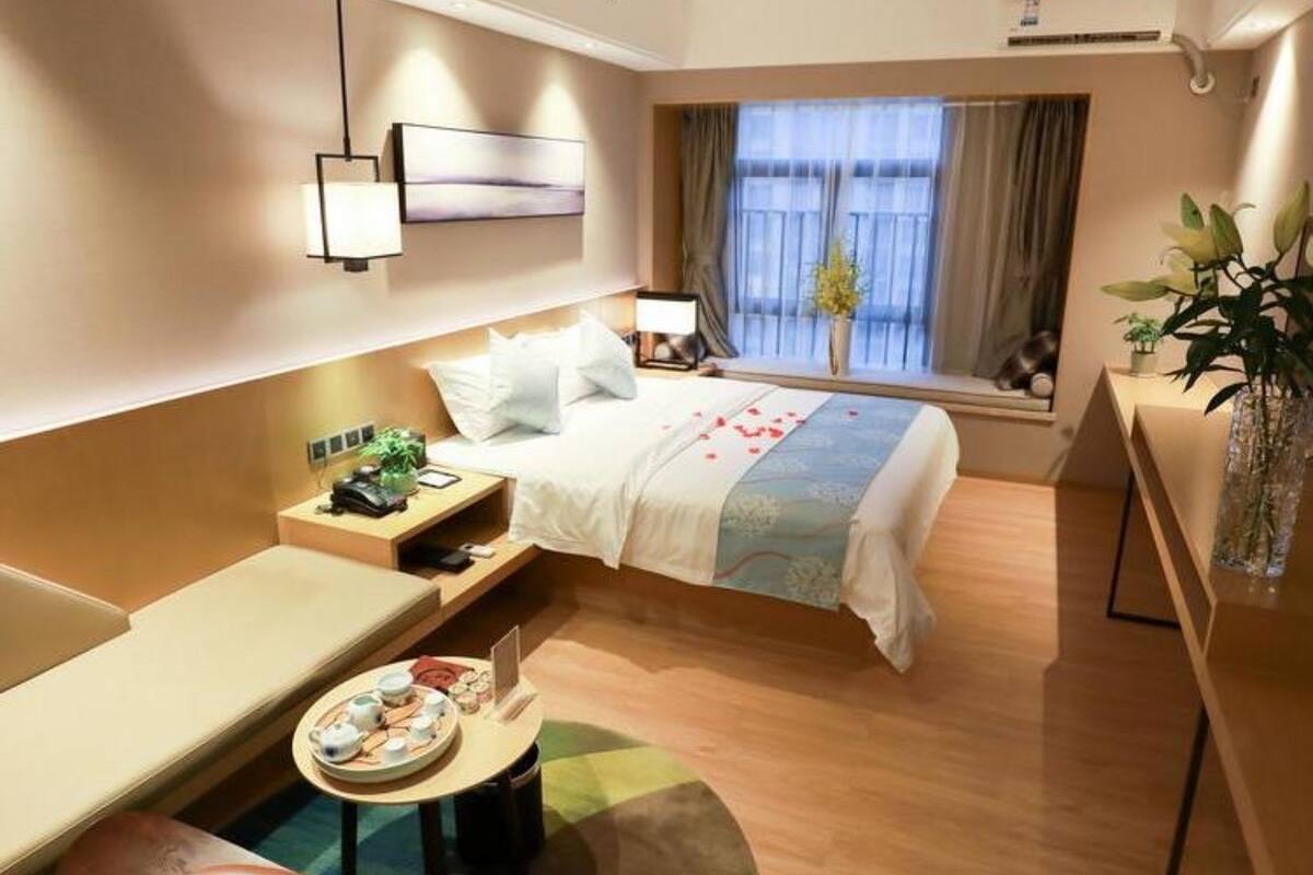 Ein Hotelzimmer mit Bett, Nachttisch, Lampe, Fenster mit Vorhängen und einer kleinen Sitzecke mit Tisch und Stühlen.