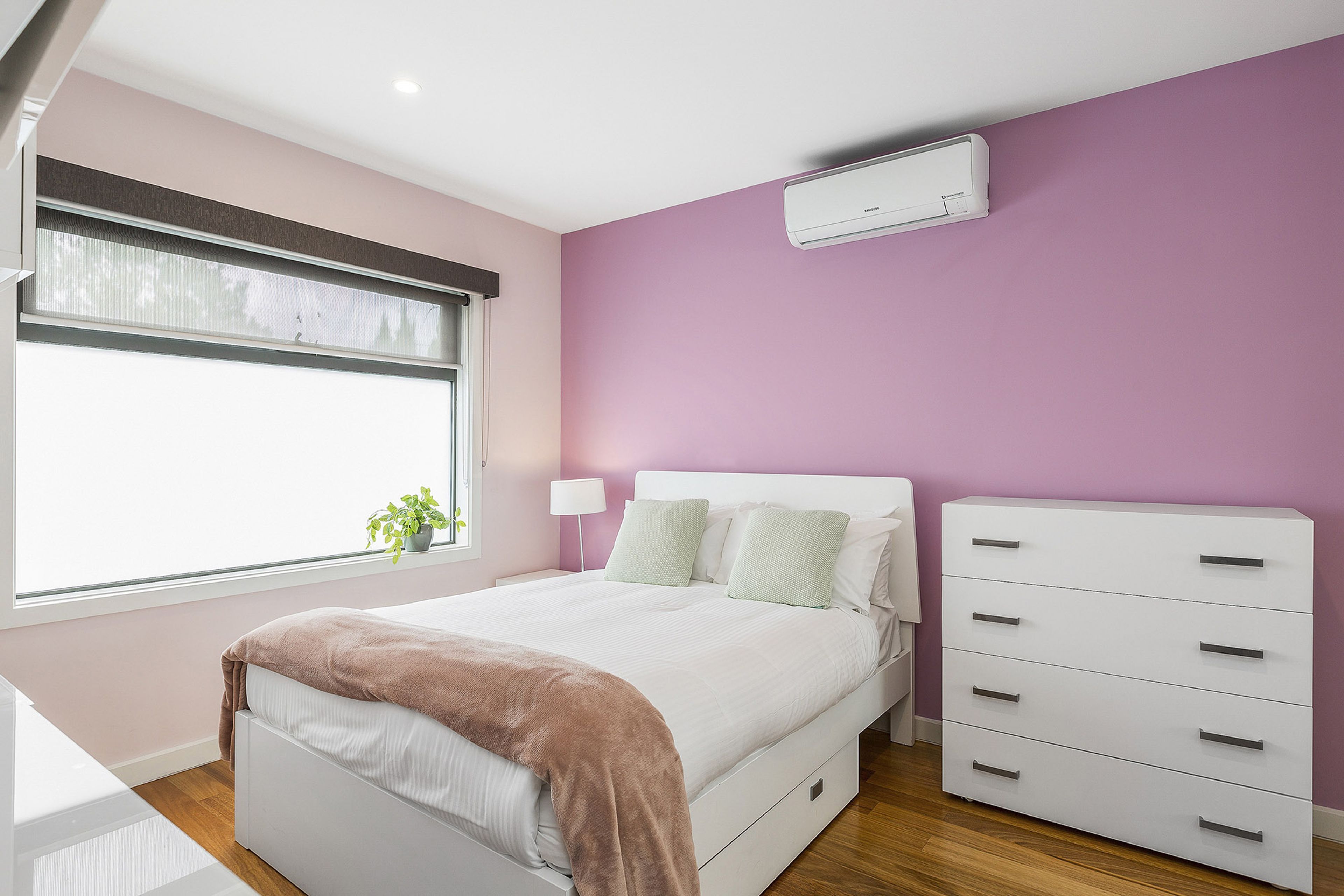 A bedroom with a bed, bedside tables, a dresser, a window with blinds, and an air conditioning unit.