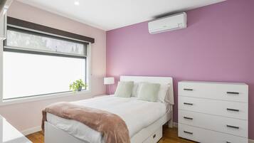 A bedroom with a bed, bedside tables, a dresser, a window with blinds, and an air conditioning unit.
