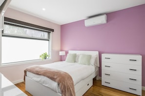 A bedroom with a bed, bedside tables, a dresser, a window with blinds, and an air conditioning unit.