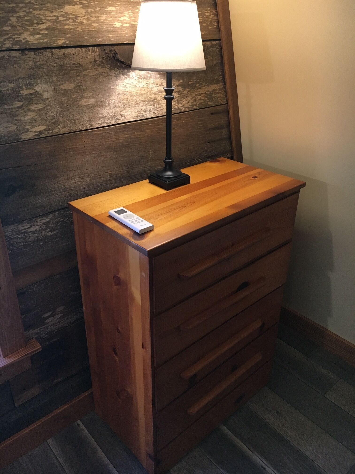 A wooden dresser with a remote control on top, a lamp on a wooden table beside it, and a wooden wall in the background.
