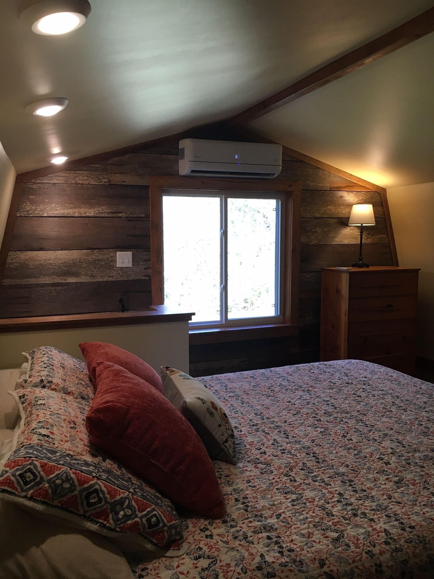 A bedroom with a sloped ceiling, wooden paneling, a large window, a bed with floral bedding and multiple pillows, a bedside table with a lamp, and a dresser.