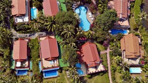 Vista aérea de un resort con varias edificaciones tipo bungaló, cada una con piscina, rodeado de palmeras y una playa.