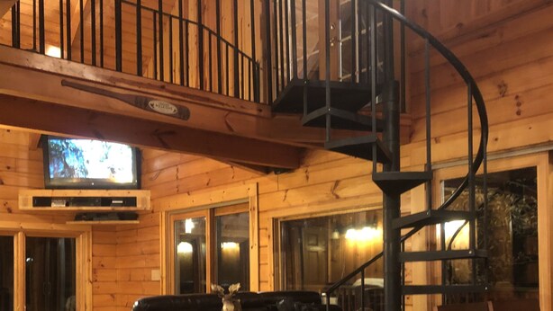 Interior de una cabaña de madera con escalera de caracol, televisor, chimenea y comedor.