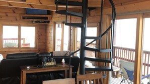 Interior de una cabaña de madera con una escalera espiral, una sala de estar con sofá y mesa, y vistas al porche exterior.
