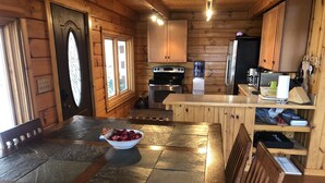 Interior de una cabaña de madera con una mesa de comedor con azulejos de piedra, un cuenco de fruta, armarios de madera, un refrigerador y una cafetera.