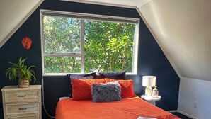 A bedroom with a large window, a bed with an orange bedspread, a nightstand with a lamp, and a small plant.