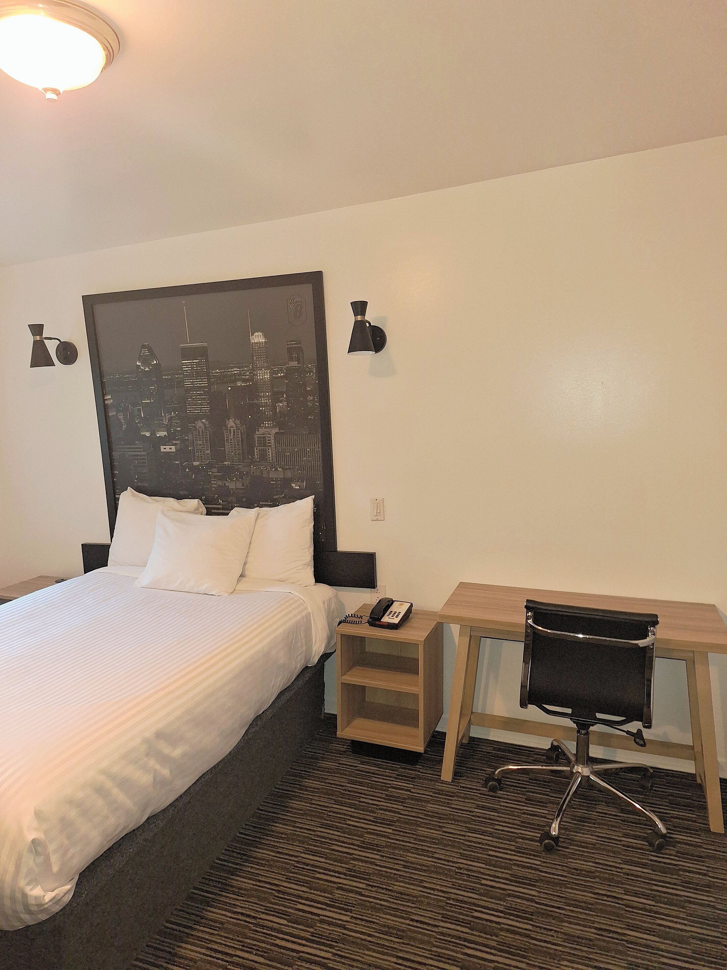 A hotel room with a bed, a desk, a chair, a nightstand, a phone, and a wall-mounted lamp.