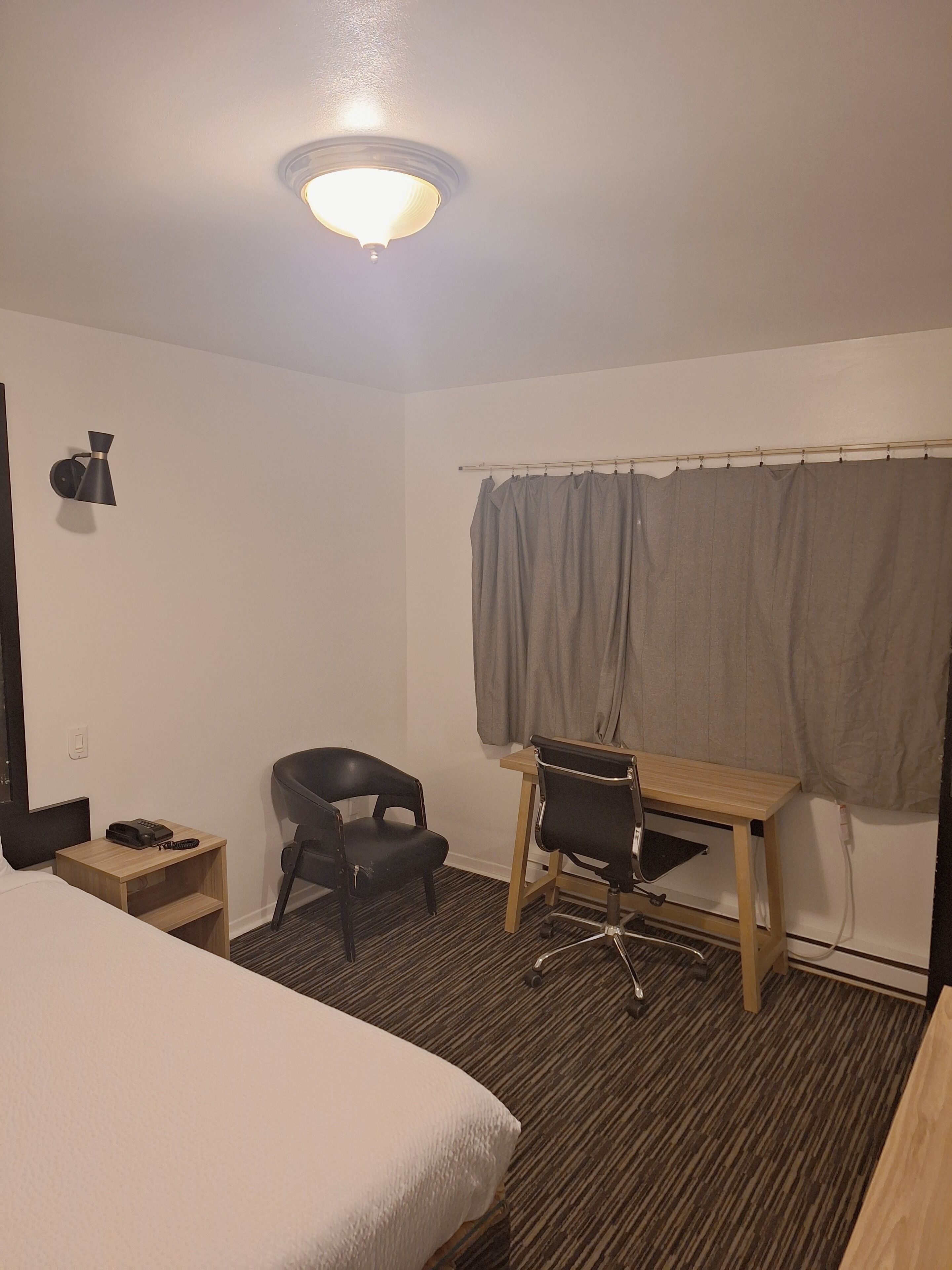 Une chambre d’hôtel comprenant un lit, un bureau, une chaise, une petite table, un téléphone et un rideau.