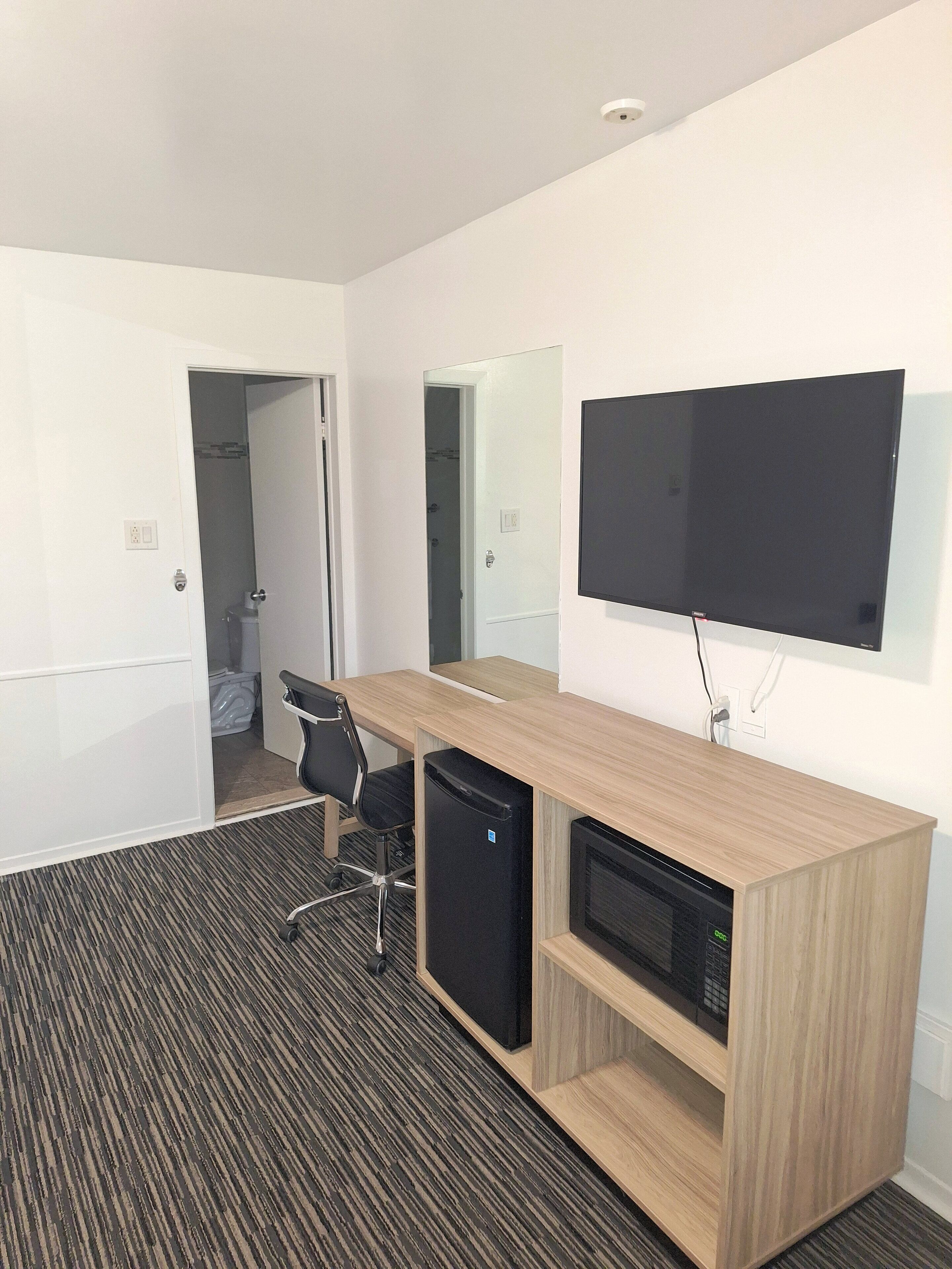 A hotel room with a wooden desk, a flat-screen TV, a mini-fridge, and a black chair.