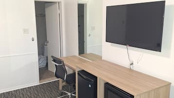 A hotel room with a wooden desk, a flat-screen TV, a mini-fridge, and a black chair.