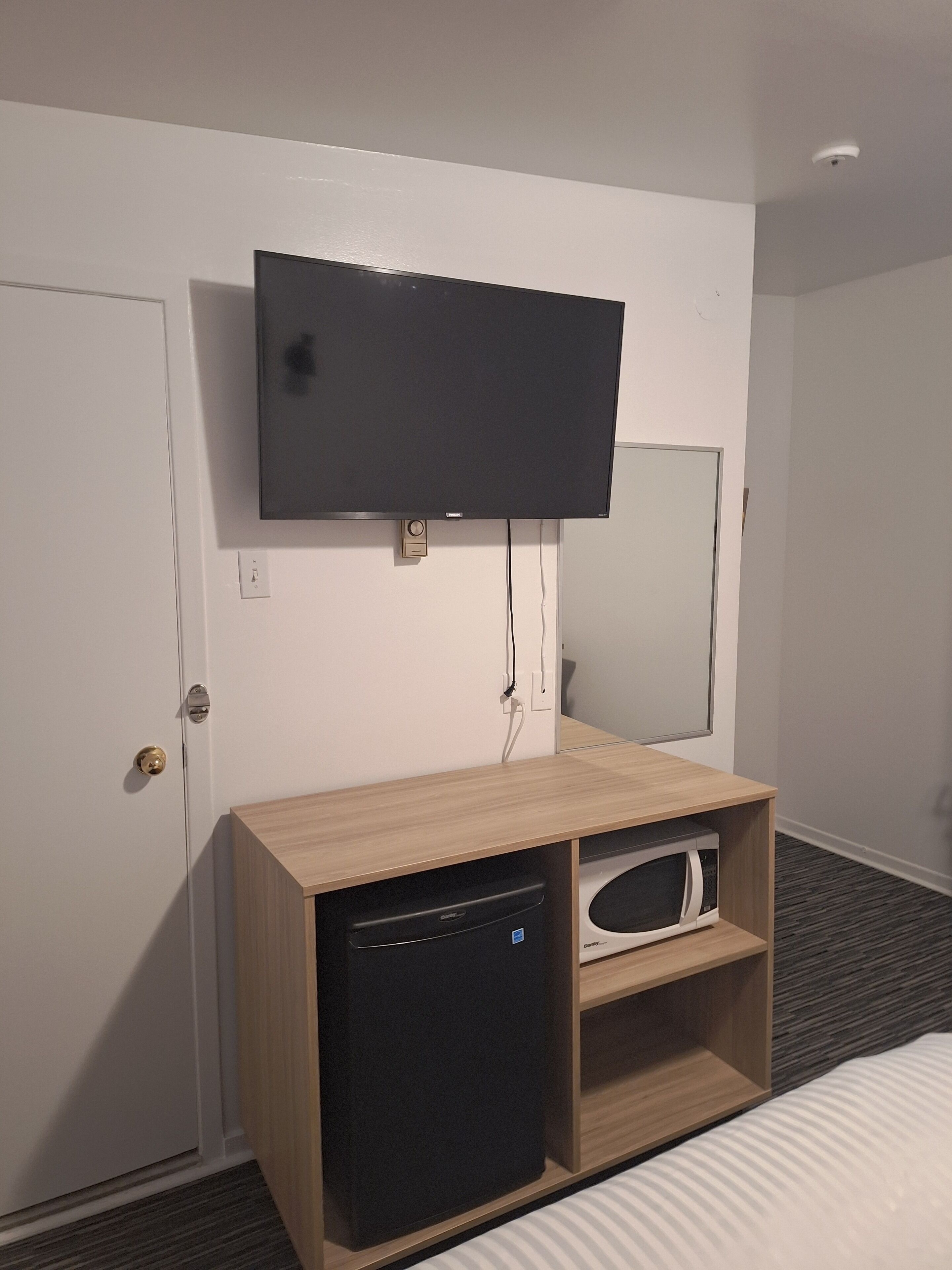 A hotel room with a flat-screen TV mounted on the wall, a wooden cabinet with a microwave, and a door with a key lock.