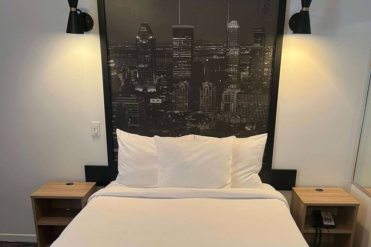 Ein Hotelzimmer mit einem Bett, zwei Nachttischen, Wandbild mit Stadtansicht und einem Telefon auf einem der Tische.