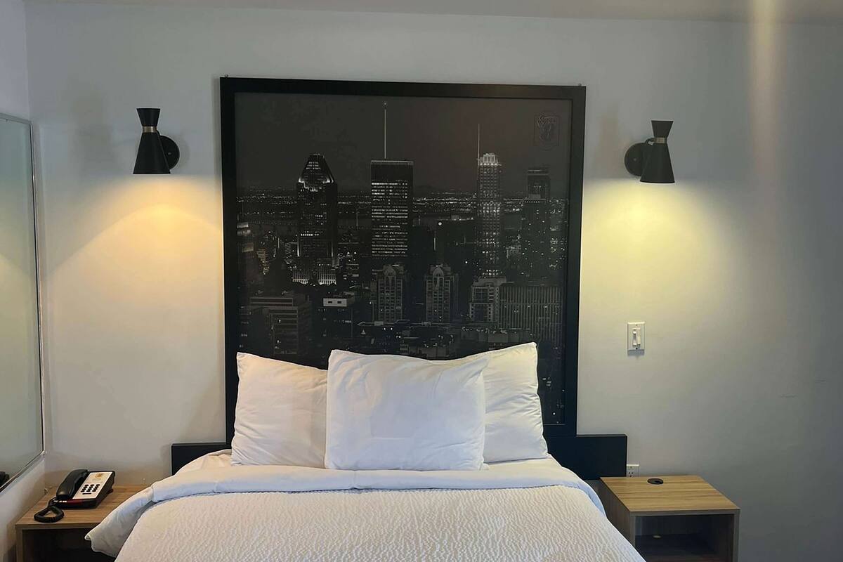 Ein Hotelzimmer mit einem Bett, weißen Kissen, einem Stadtbildgemälde an der Wand und zwei Wandleuchten.
