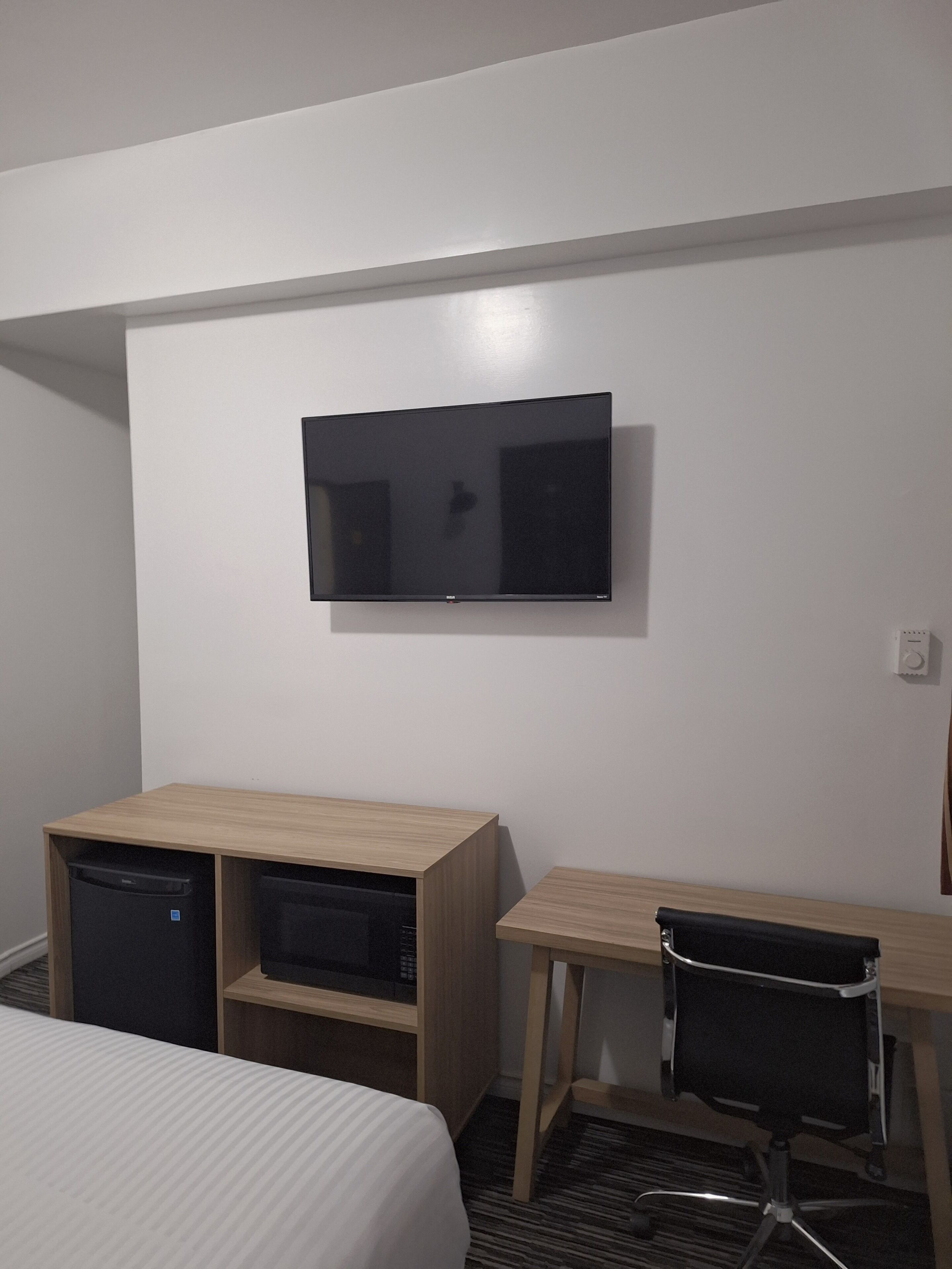 Una camera d'albergo con un letto, una scrivania con una sedia, una televisione a parete e un piccolo frigorifero.