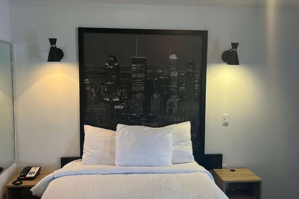 Une chambre d’hôtel avec un lit, des oreillers blancs, une œuvre d’art représentant un paysage urbain accrochée au mur et deux lampes de chevet.