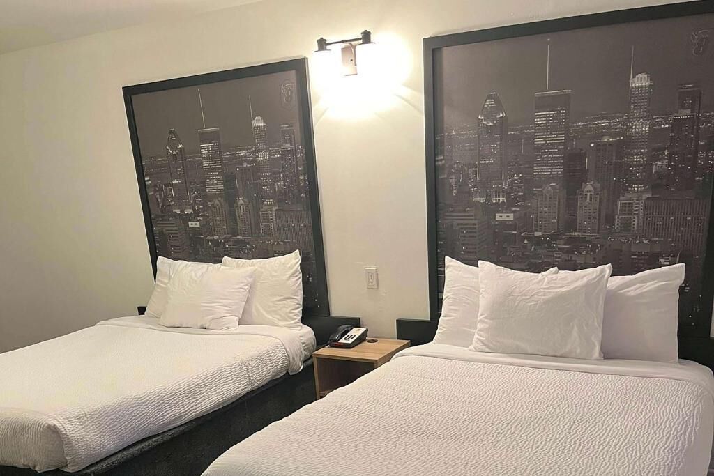 Habitación de hotel con dos camas, cada una con ropa de cama y almohadas blancas. Hay una mesita de noche con un teléfono y una lámpara entre las camas. Las paredes tienen grandes cuadros enmarcados con vistas a un horizonte urbano.