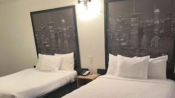 Quarto de hotel com duas camas, cada uma com roupa de cama e almofadas brancas. Há uma mesinha de cabeceira com telefone e abajur entre as camas. As paredes apresentam grandes quadros emoldurados de um horizonte urbano.