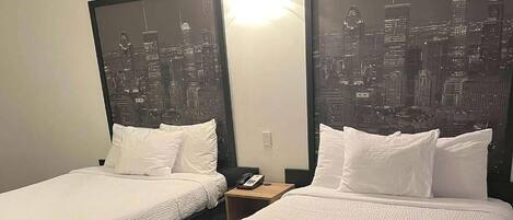 Habitación de hotel con dos camas, cada una con ropa de cama y almohadas blancas. Hay una mesita de noche con un teléfono y una lámpara entre las camas. Las paredes tienen grandes cuadros enmarcados con vistas a un horizonte urbano.