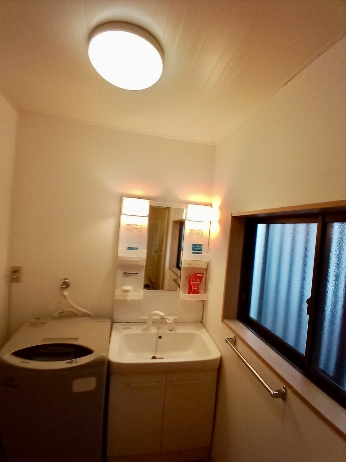 Un baño pequeño con inodoro, lavamanos y espejo. Tiene una ventana con cortinas, un tendedero para toallas y un dispensador de jabón.
