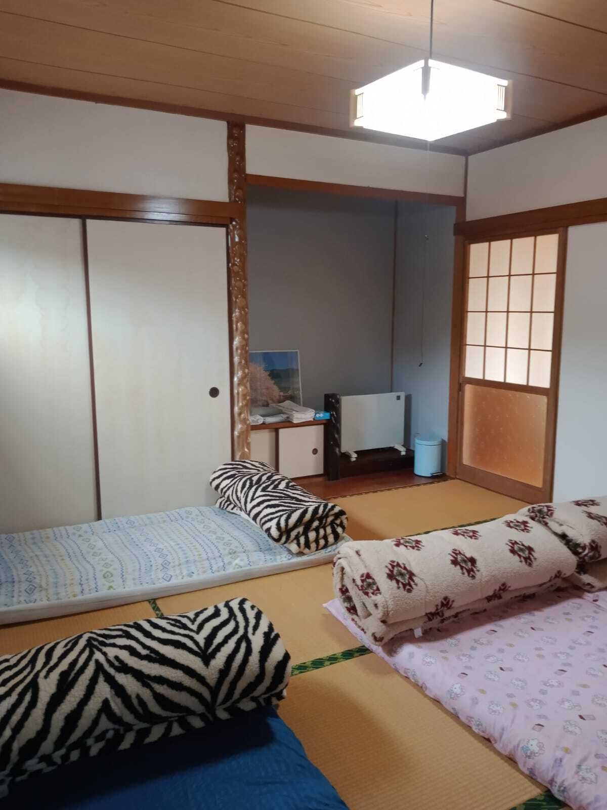 A traditional Japanese-style room with tatami flooring, a futon bed with patterned bedding, a small desk with a chair, and a sliding door.