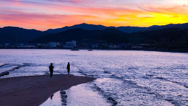 Una playa con dos personas de pie sobre la arena, un mar tranquilo y una puesta de sol sobre montañas.