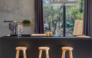 A modern kitchen with a large window, wooden stools, and a plant on the counter. - Gorgeous home in Katuni (Katuni)