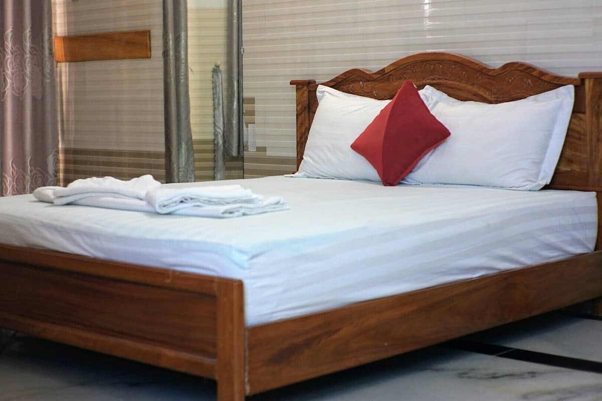 Uma cama bem arrumada com lençóis brancos e um travesseiro vermelho, uma cabeceira de madeira e uma porta de vidro ao fundo.