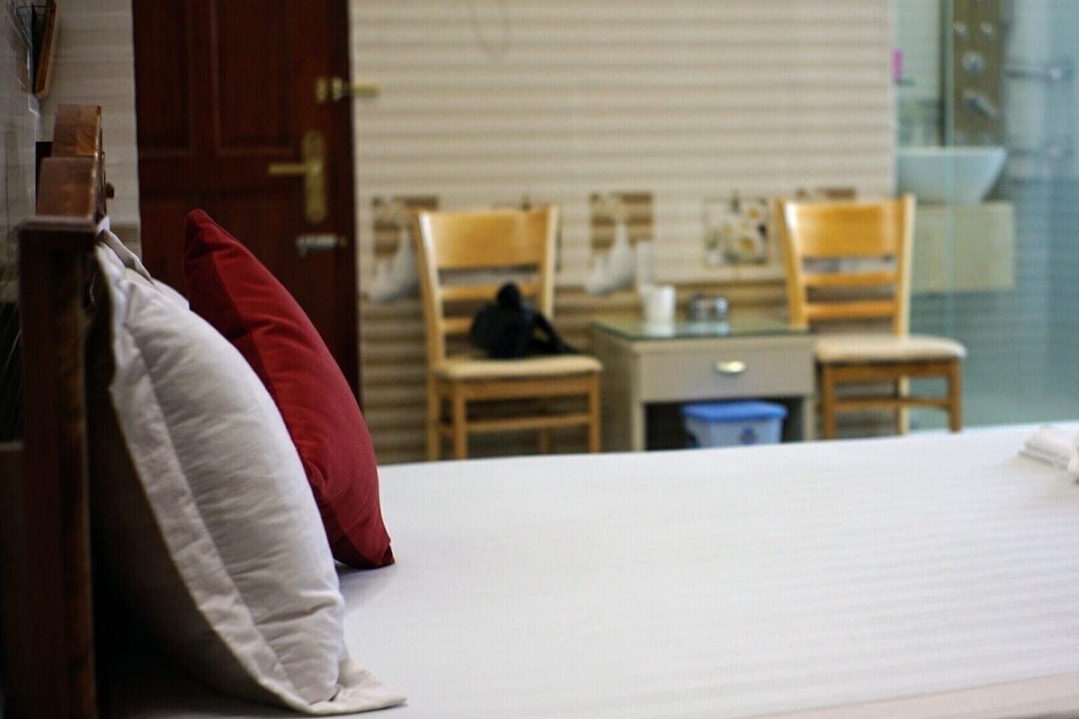 Quarto de hotel com cama, escrivaninha, duas cadeiras e janela com persianas.