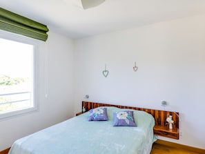 Een slaapkamer met een bed, een raam met een groene gordijn, een houten hoofdbord en een wandtafeltje met een vaas.