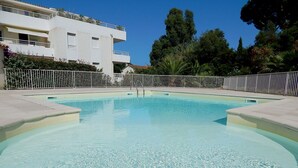 Una piscina con superficie de agua azul cristalina, rodeada por una cerca blanca y adyacente a un edificio blanco con balcones.